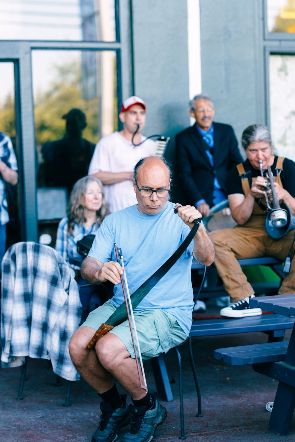 Bald man with glasses plays a unique stringed instrument outdoors while musicians and audience gather behind him.