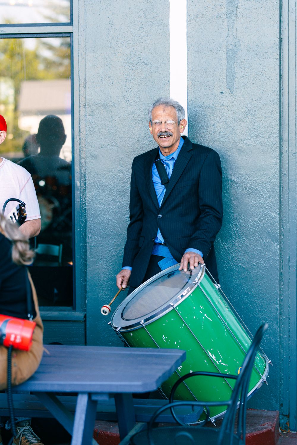 Smiling elderly man in a black suit holding a green drum with drumsticks outdoors against a blue wall.