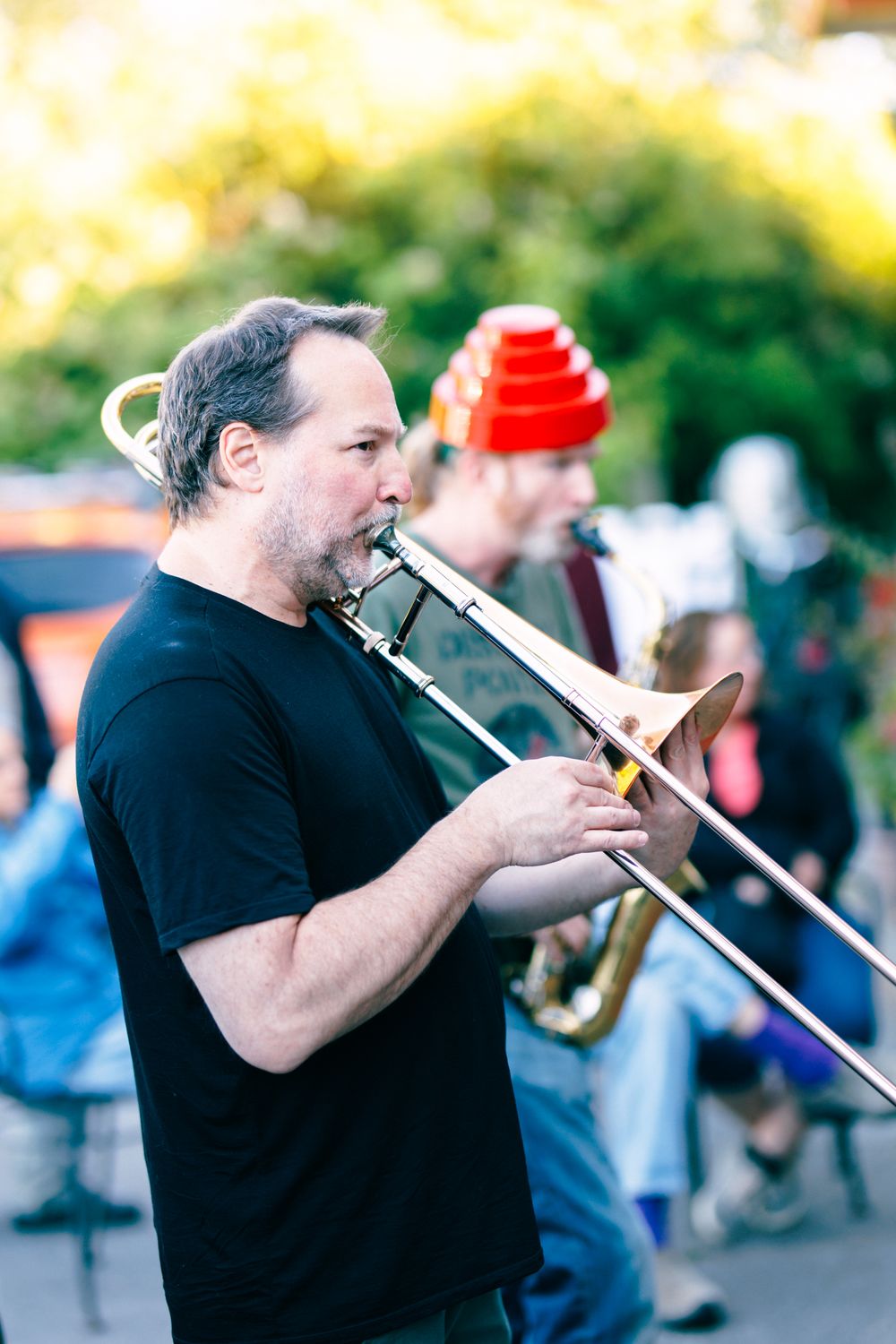 A man in a black t-shirt plays trombone outdoors at a street music performance with other musicians behind him.