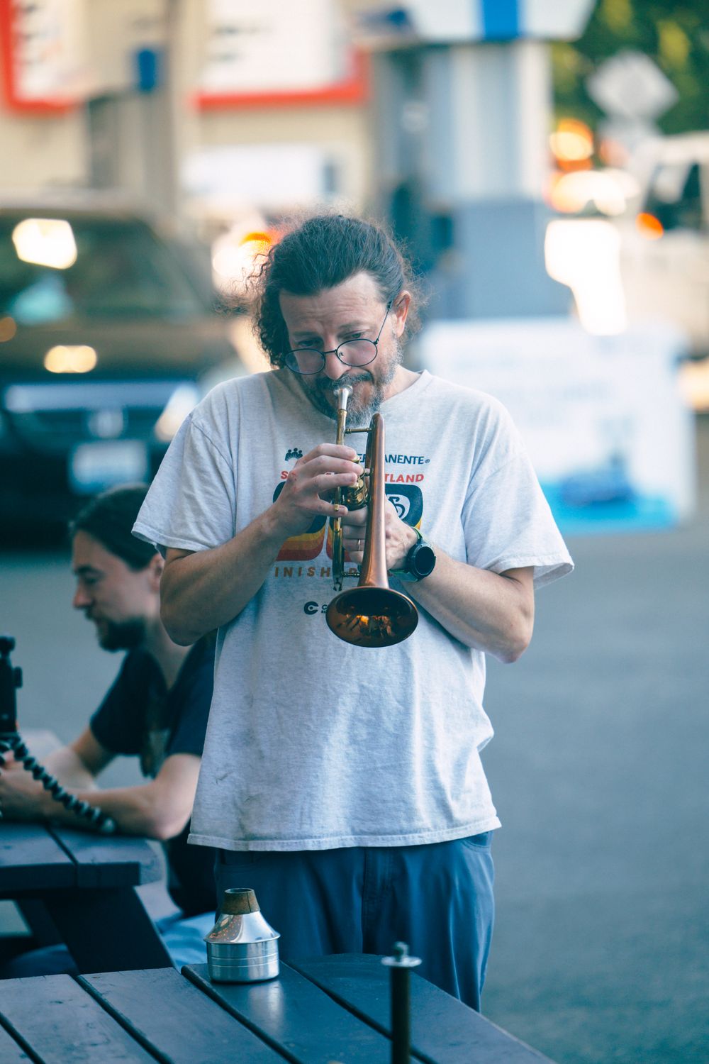 Long-haired man in white t-shirt plays trumpet outdoors at a table on a city street.