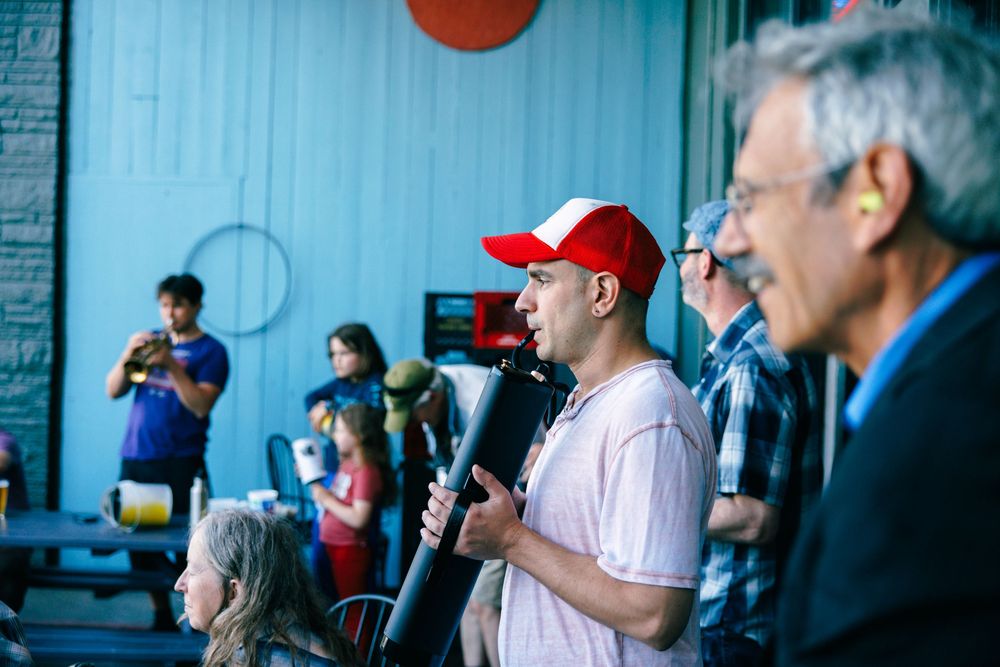 A man in a red cap speaks into a microphone at an indoor event with an audience and colorful backdrop behind him.