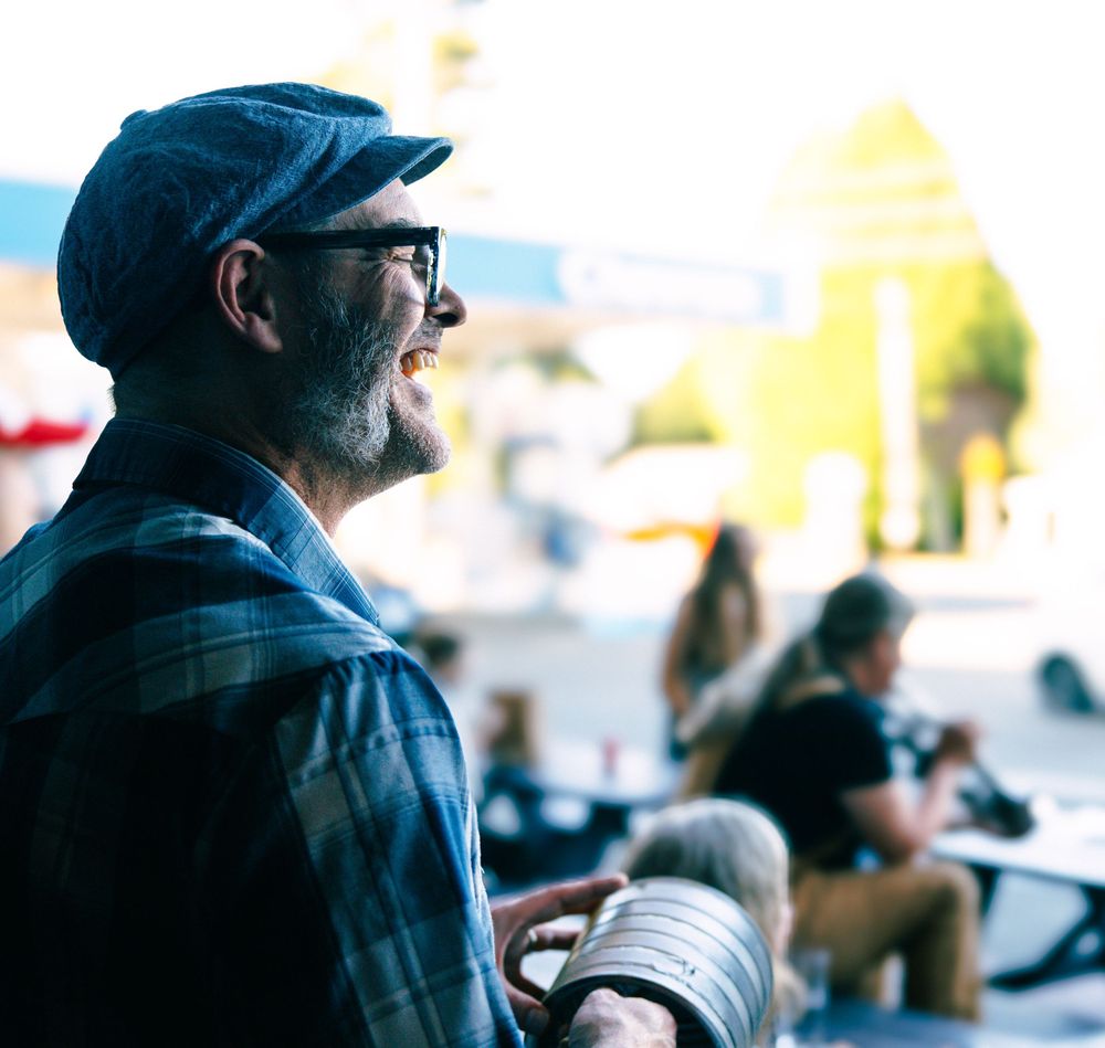 Smiling bearded man wearing glasses and a flat cap enjoys an outdoor cafe setting in bright sunlight.