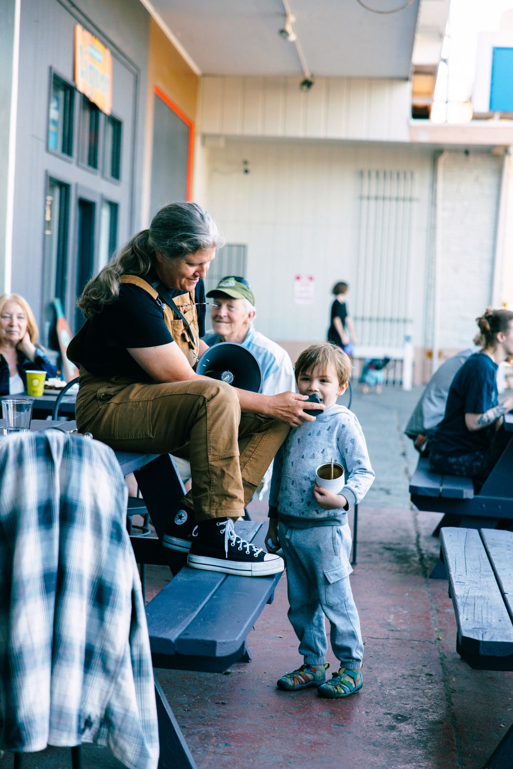 A long-haired adult and elderly man interact with a young child holding a drink at an outdoor venue with picnic tables.