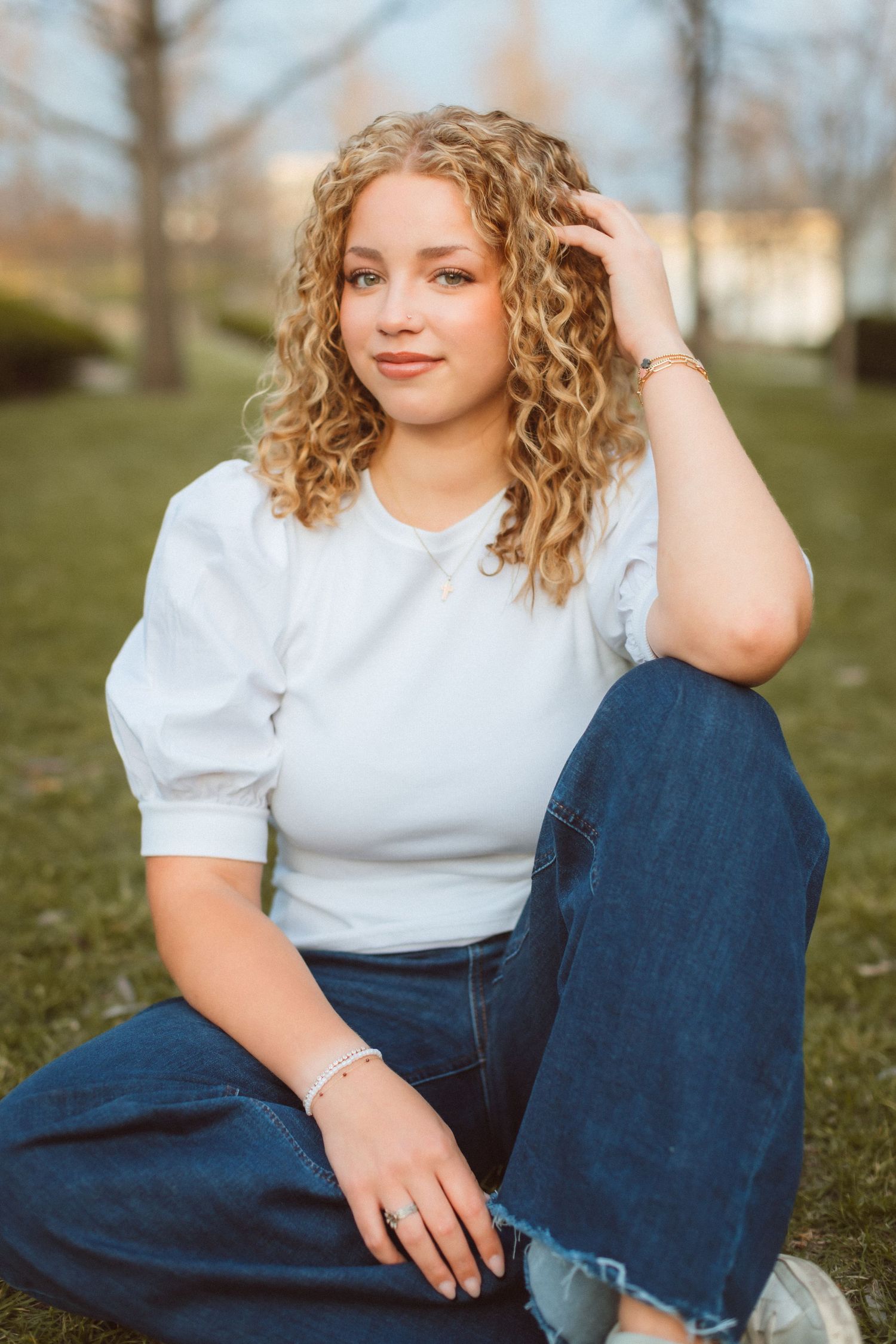 A young woman sits with one knee up, resting her elbow on her knee and tousling her hair with one hand as she smiles softly at the camera.