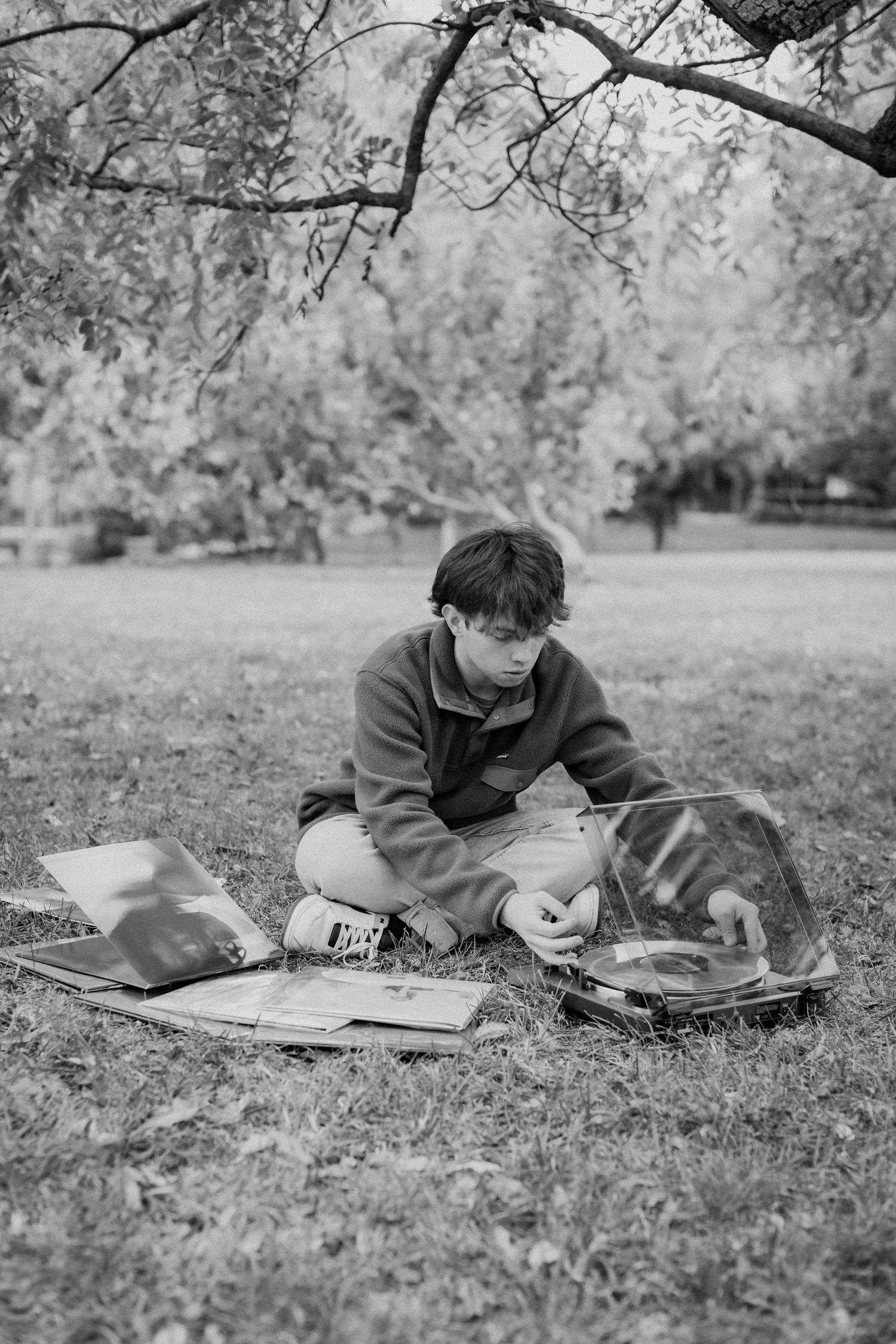 A senior guy sits cross-legged on the grass in a park with his vinyl record collection spread out in front of him.