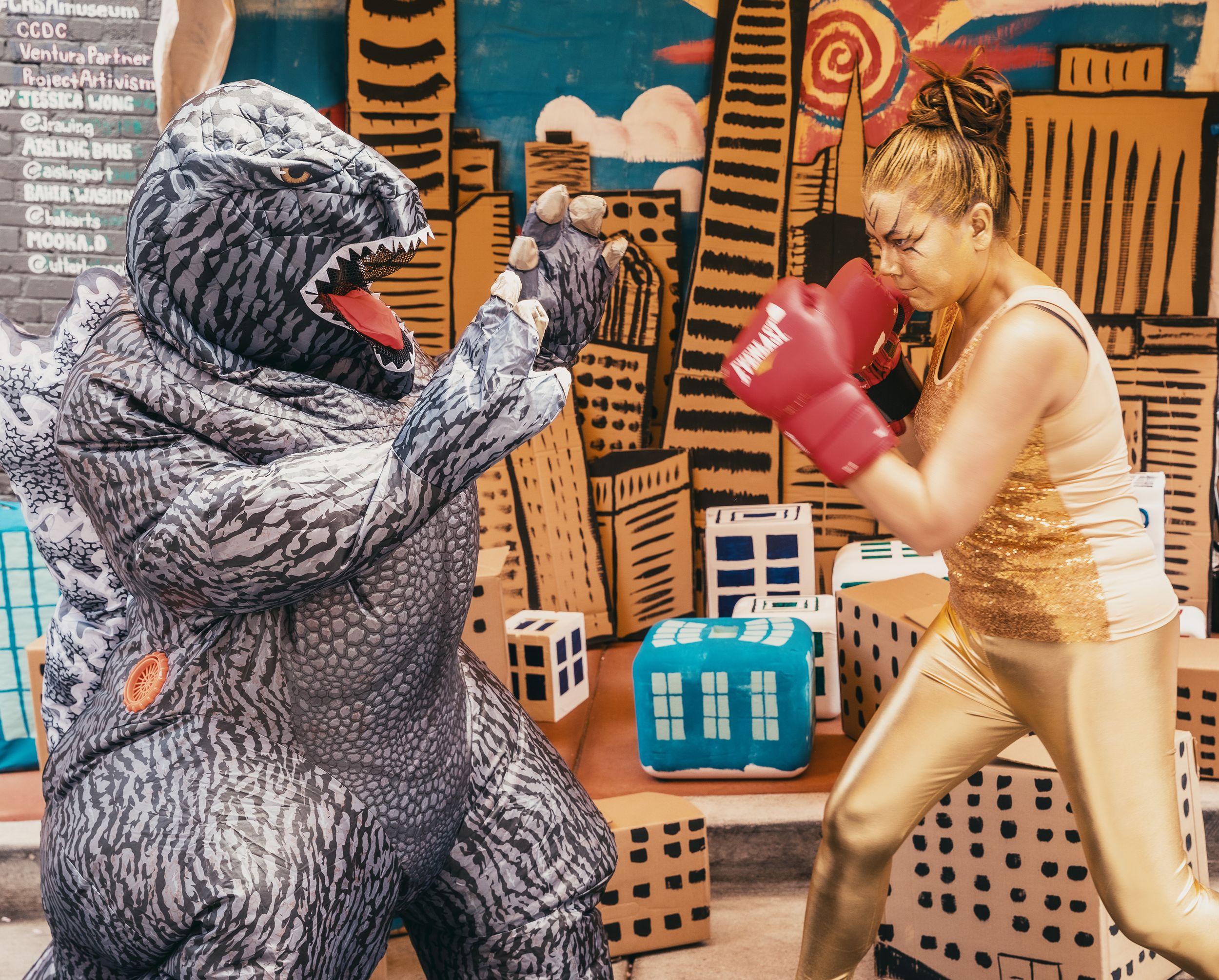 Woman in boxing gloves sparring with a Godzilla costume character in front of a colorful city mural backdrop.