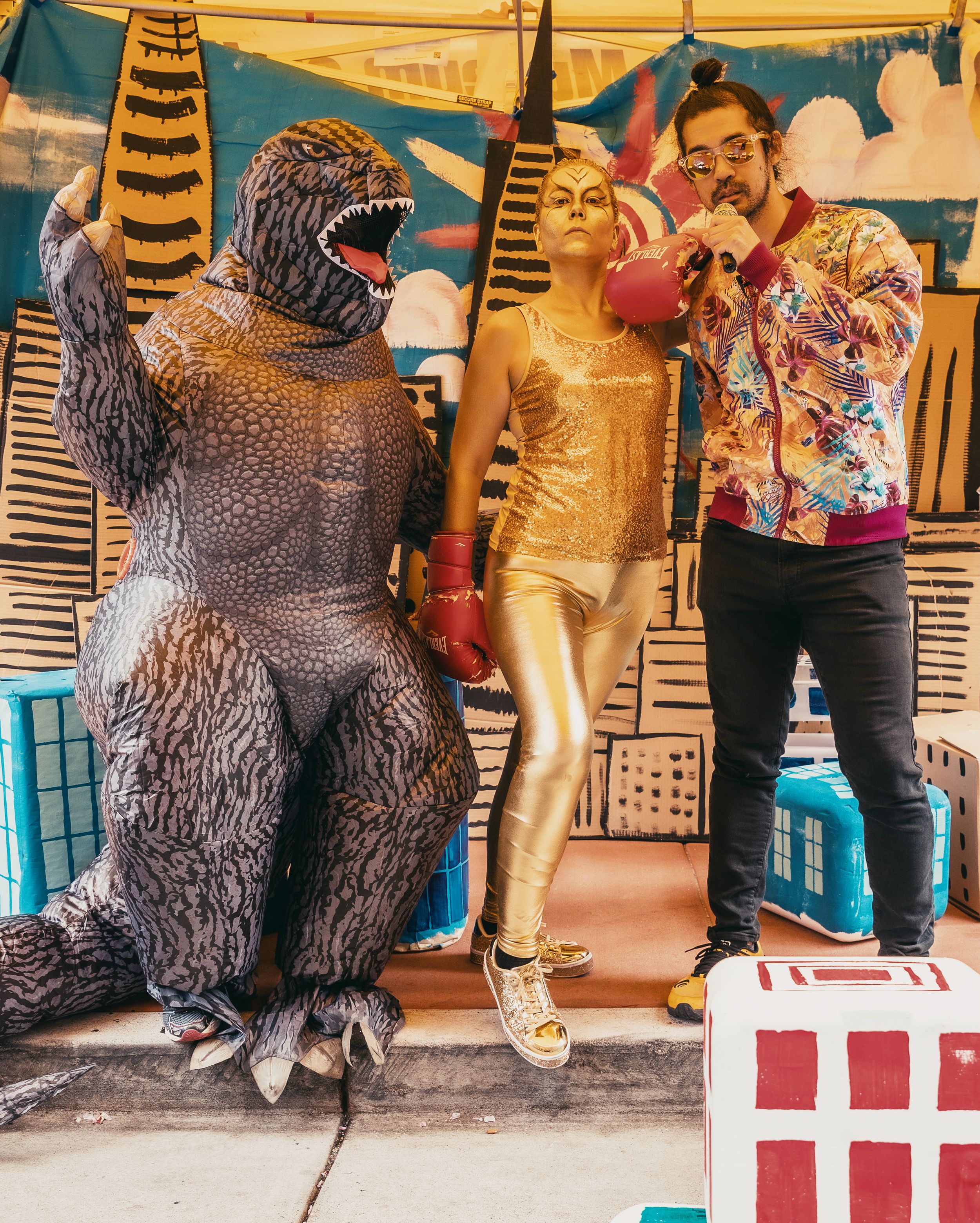 Two people in gold and floral costumes pose with a Godzilla inflatable suit in front of a colorful city mural backdrop.