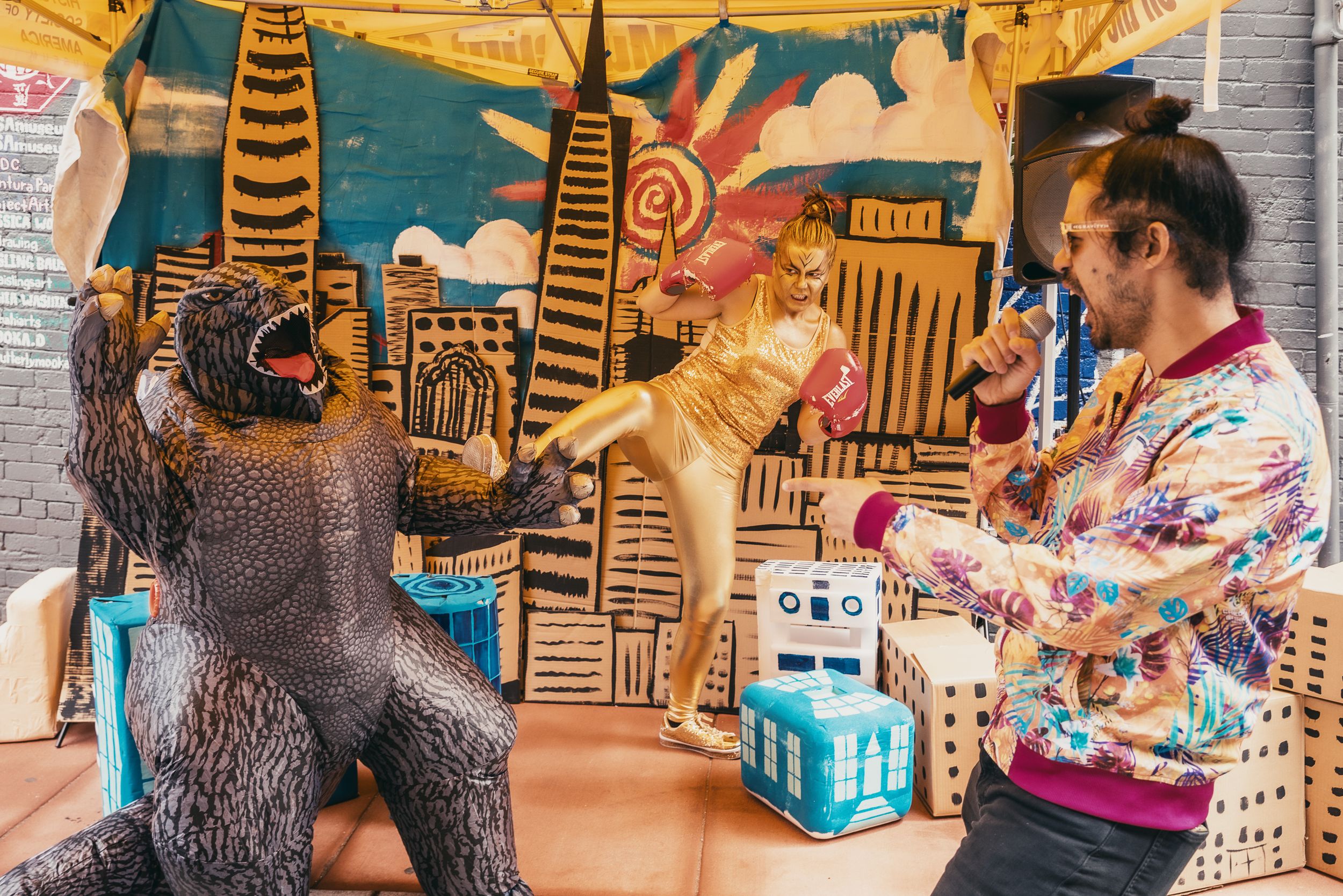 Man in colorful shirt poses with gorilla and dinosaur costumes against a painted cityscape backdrop.
