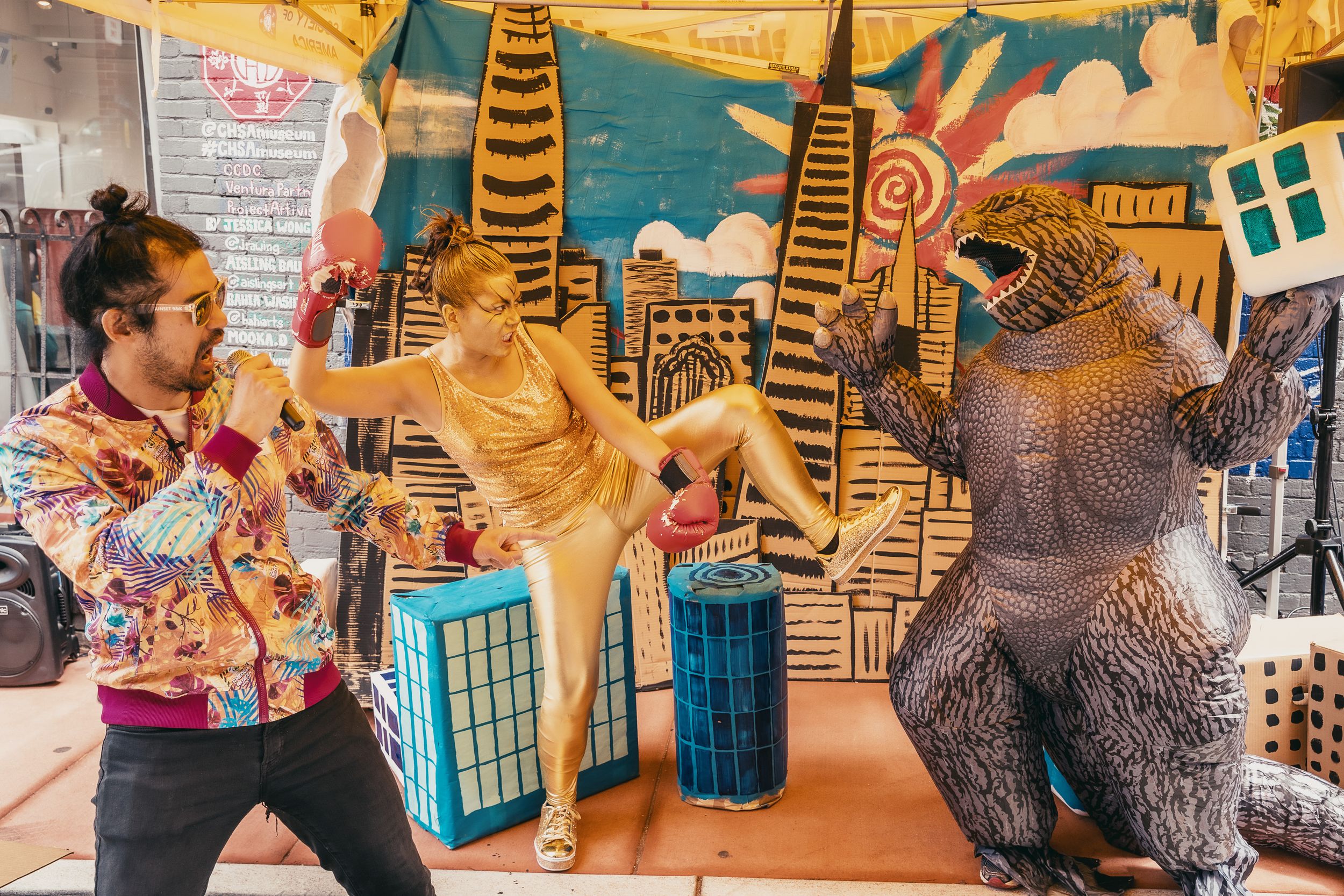 Two people pose with a Godzilla costume in front of a colorful city mural backdrop.