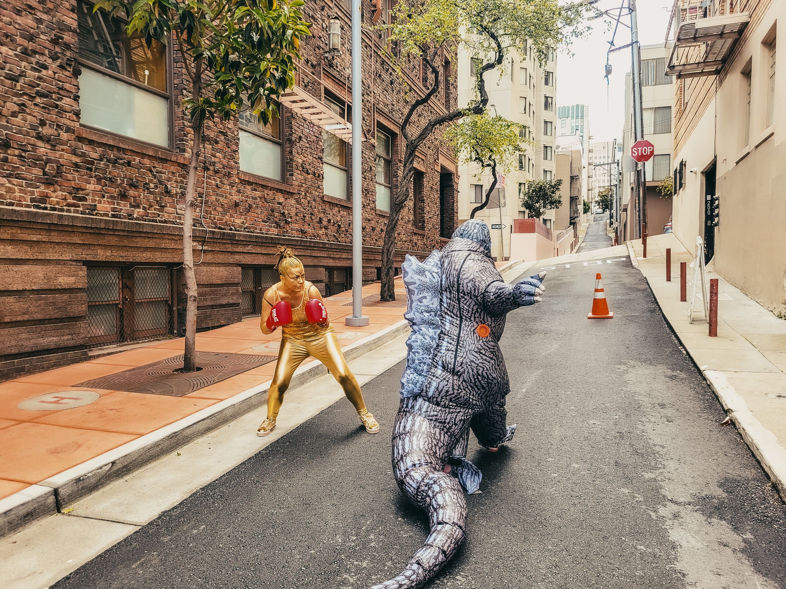 Person in gold boxing costume faces inflatable T-Rex dinosaur costume on urban city street in playful confrontation.
