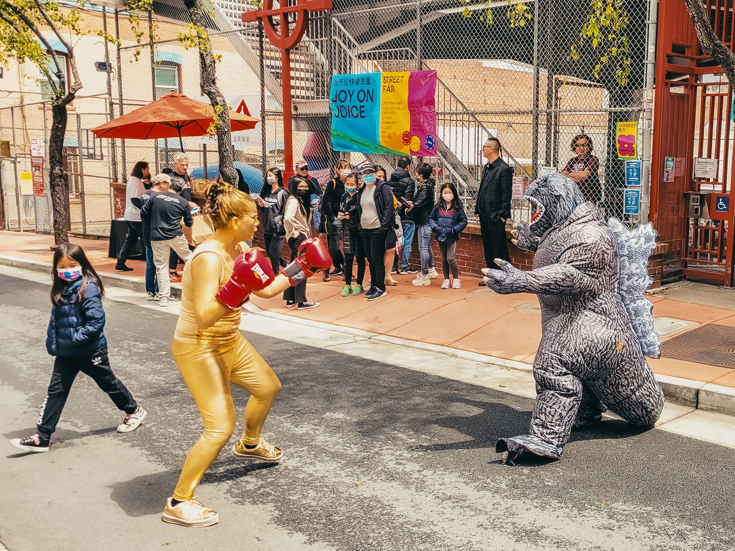 Street performers in costumes, one dressed as a lion and another as a gorilla, mock-boxing on a city sidewalk.