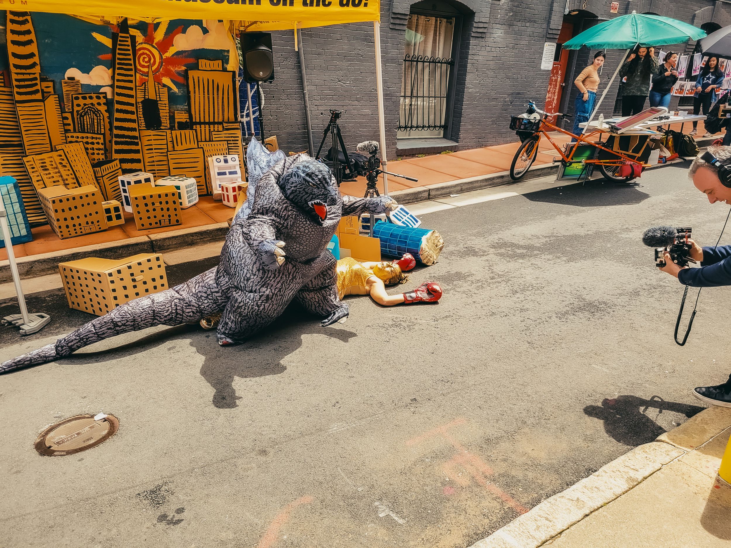 Person in Godzilla costume performing street theater act on urban pavement with colorful city mural backdrop.