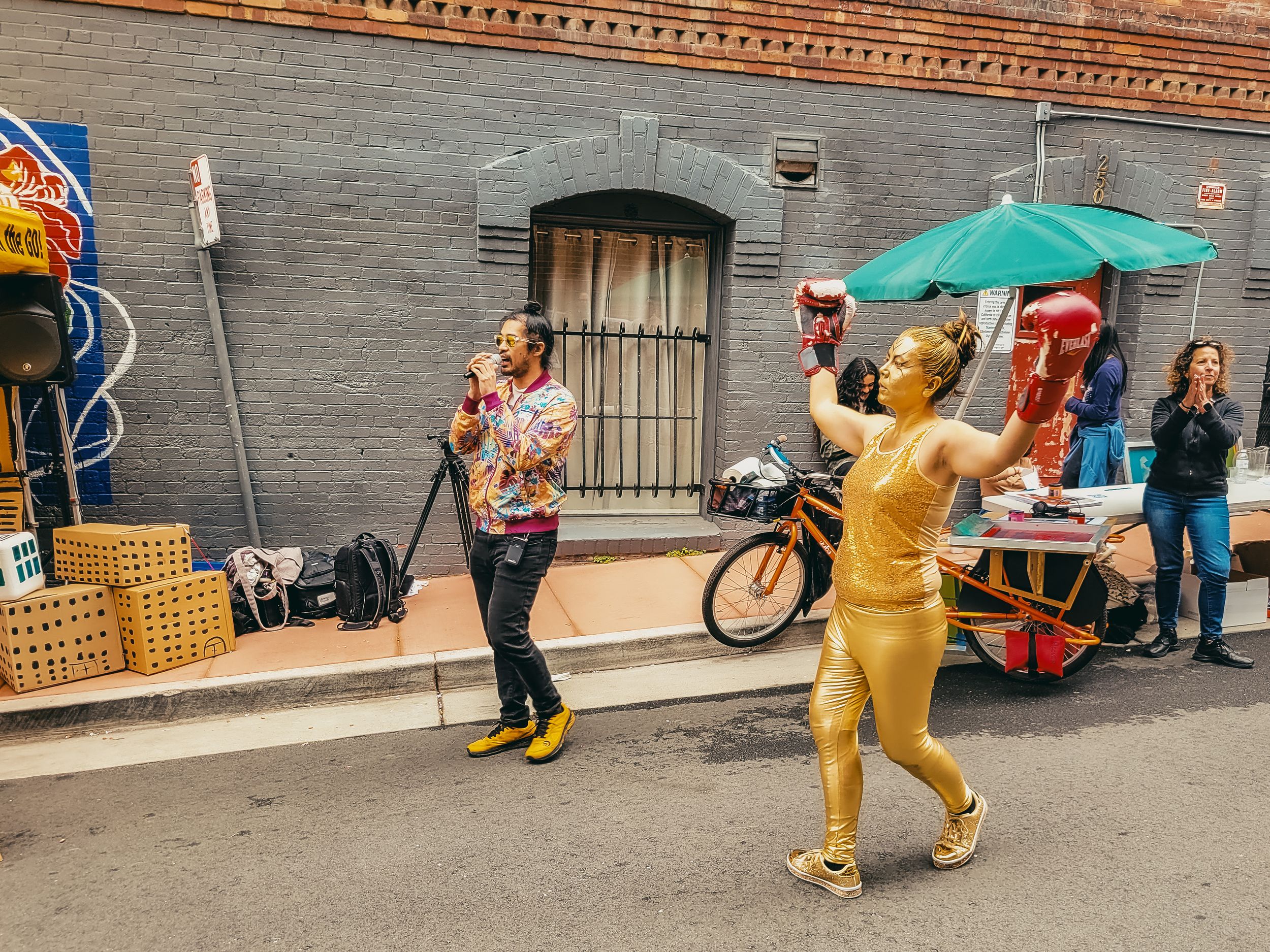 Gold-painted street performer poses near a food cart while a man in colorful shirt watches on a city alley.