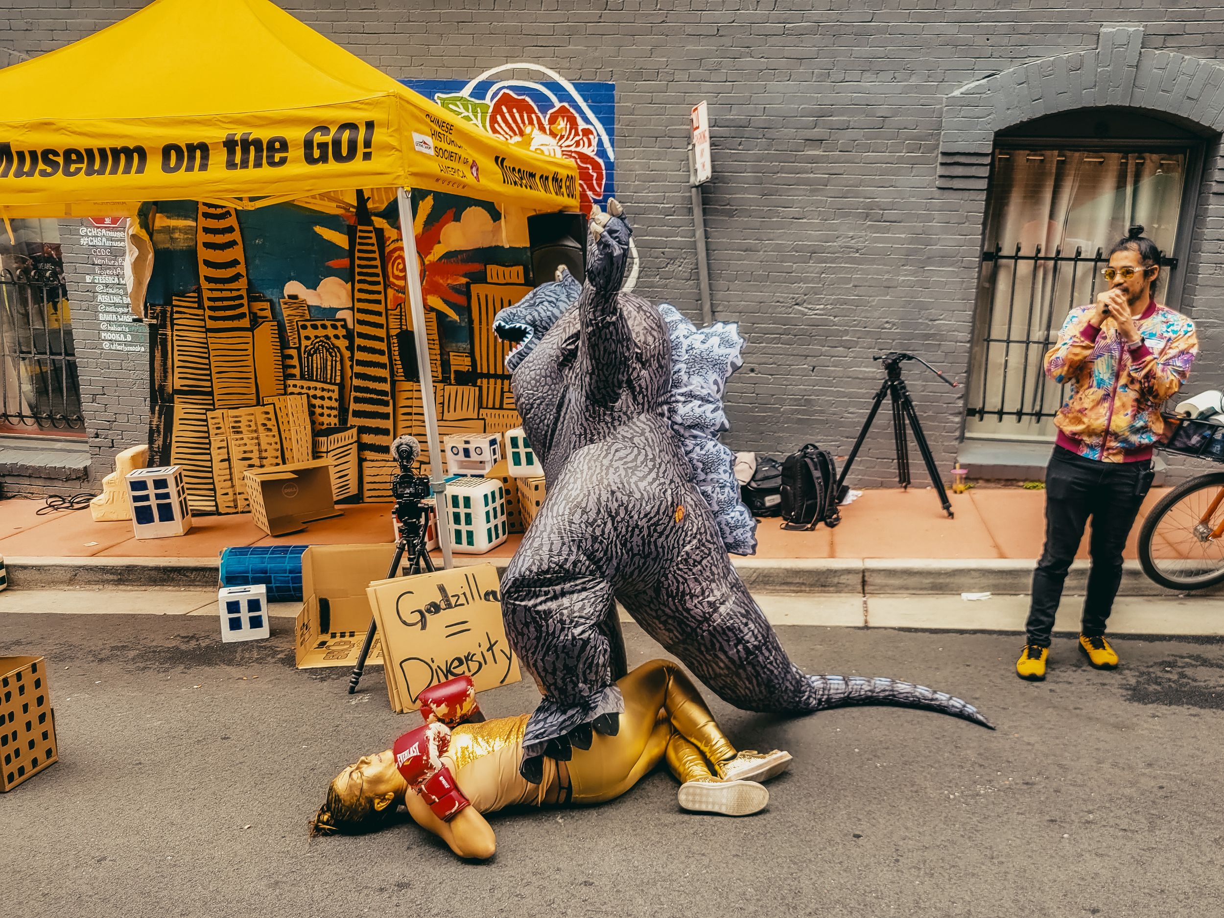 Person in Godzilla costume attacks inflatable hot dog near Museum on the GO tent at outdoor street festival.