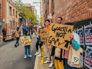 People holding Godzilla protest signs on a city street, wearing yellow outfits during an outdoor demonstration.
