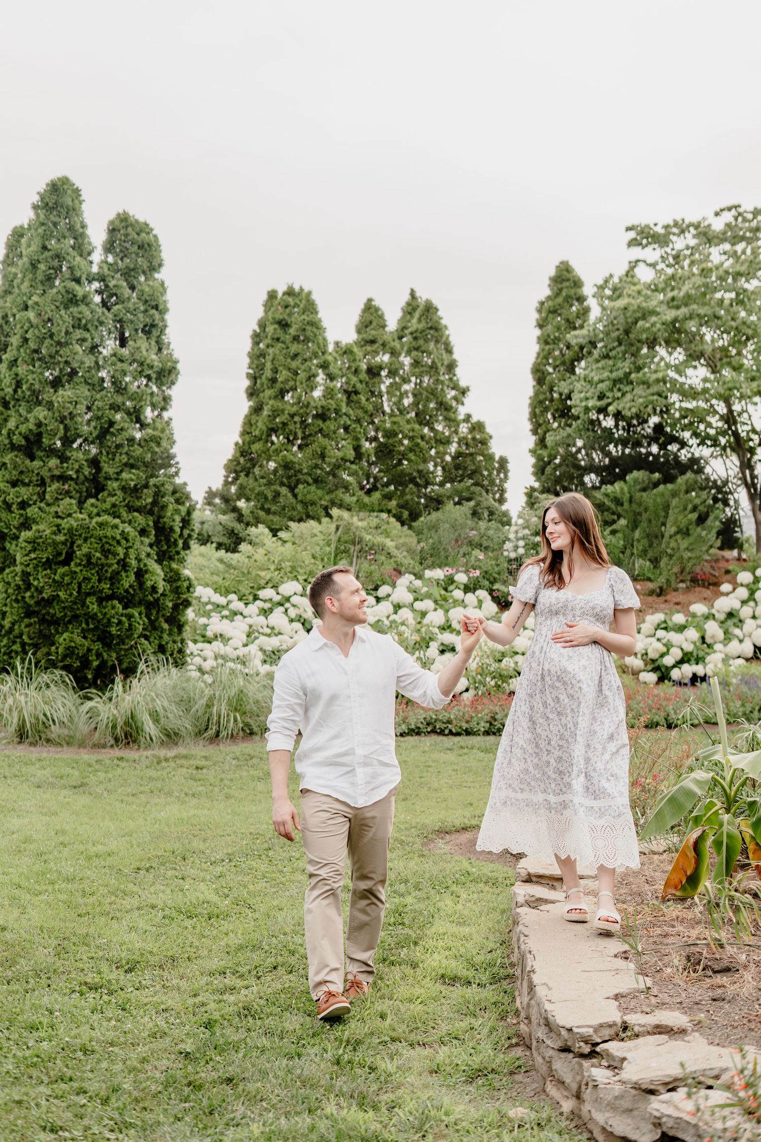 A pregnant couple holds hands while walking through a lush garden with white flowers and tall trees.