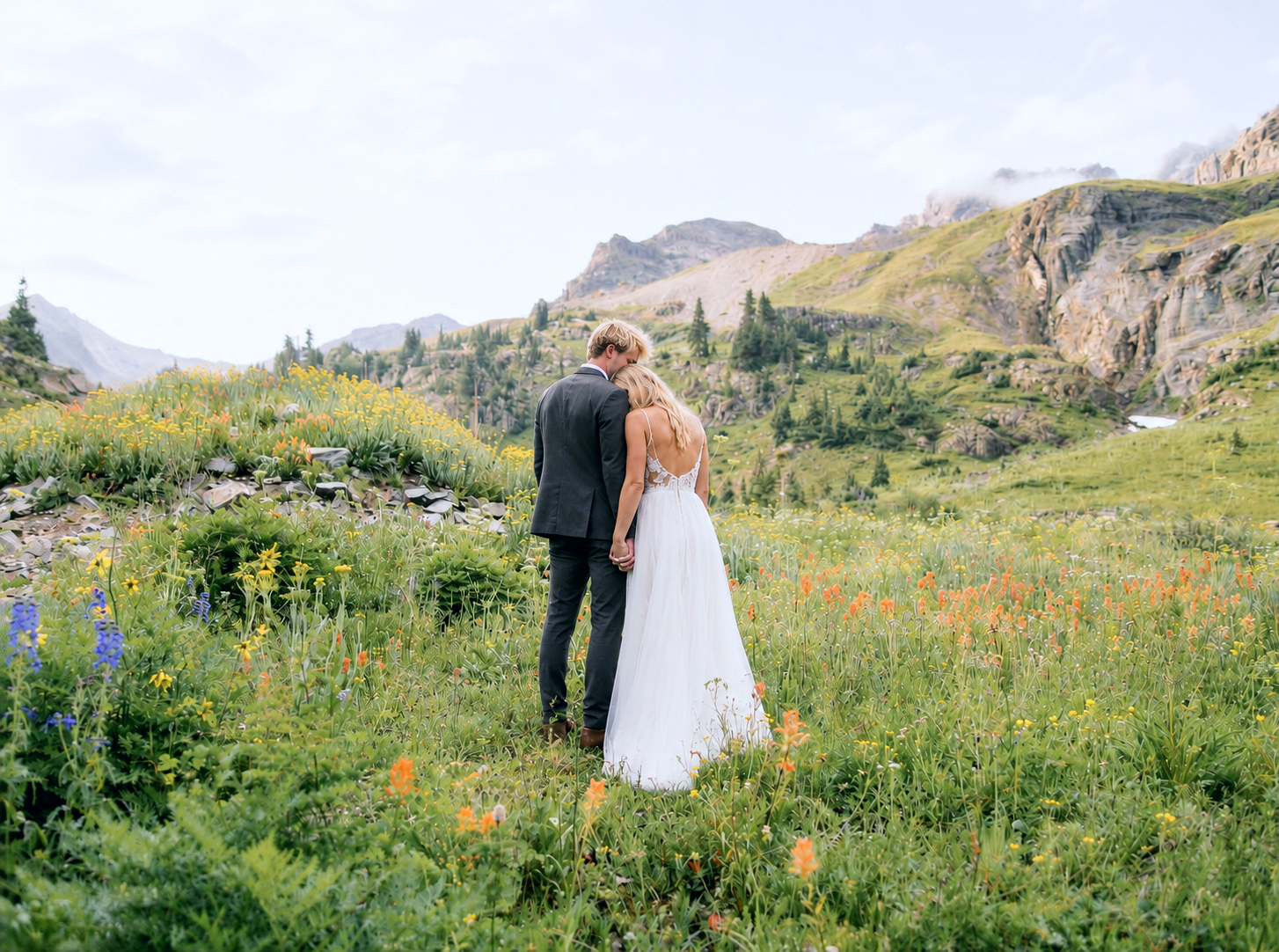 Bride and groom embrace near a mountain lake with evergreen trees, waterfall, and snow-capped peaks in Colorado.