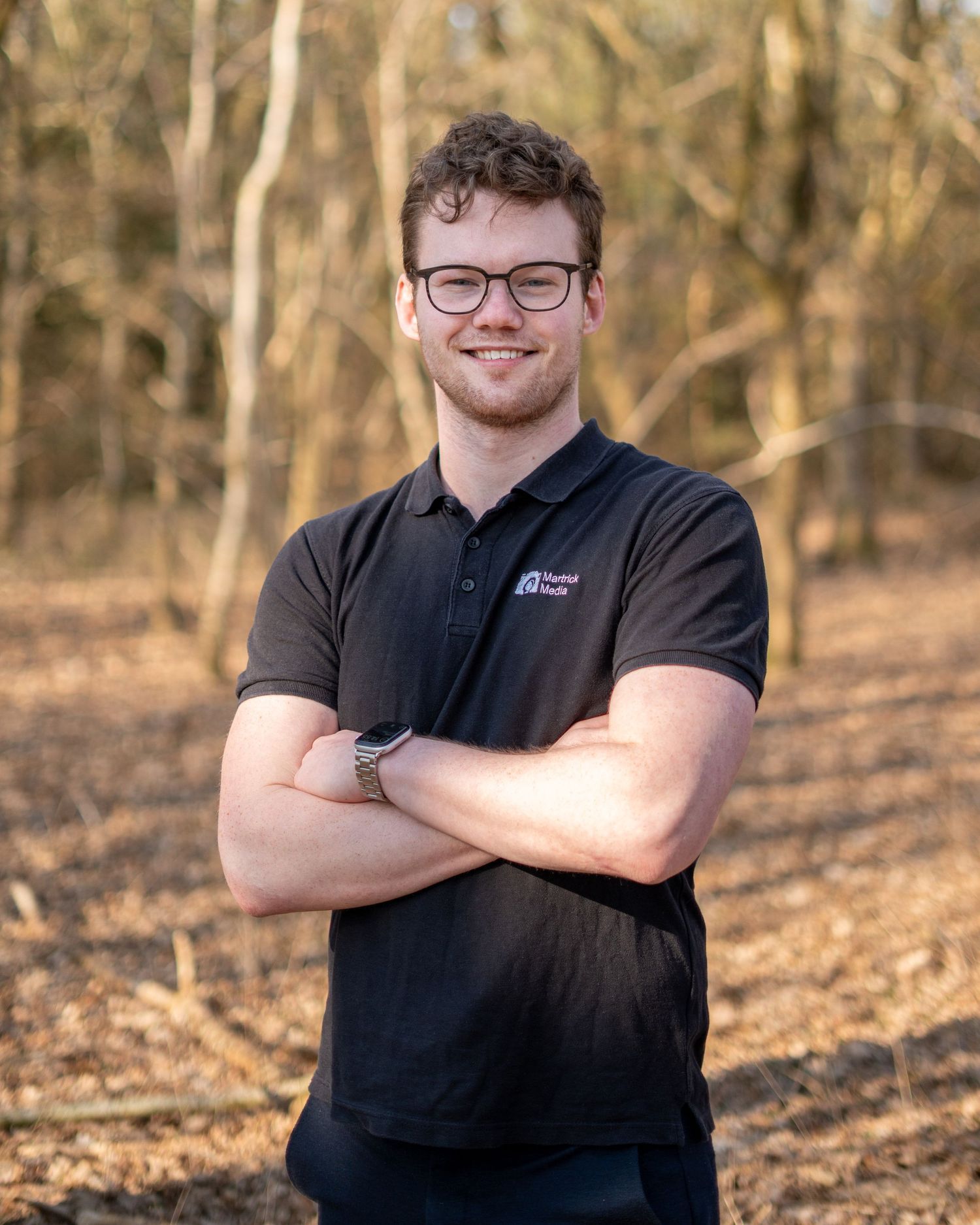Young man with glasses and crossed arms smiling outdoors in a black polo shirt among autumn trees.