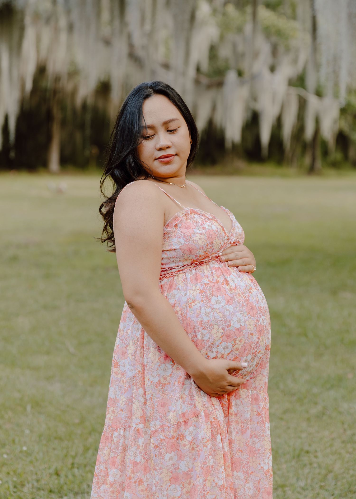 Pregnant woman in pink floral maxi dress cradling her baby bump outdoors under Spanish moss trees.