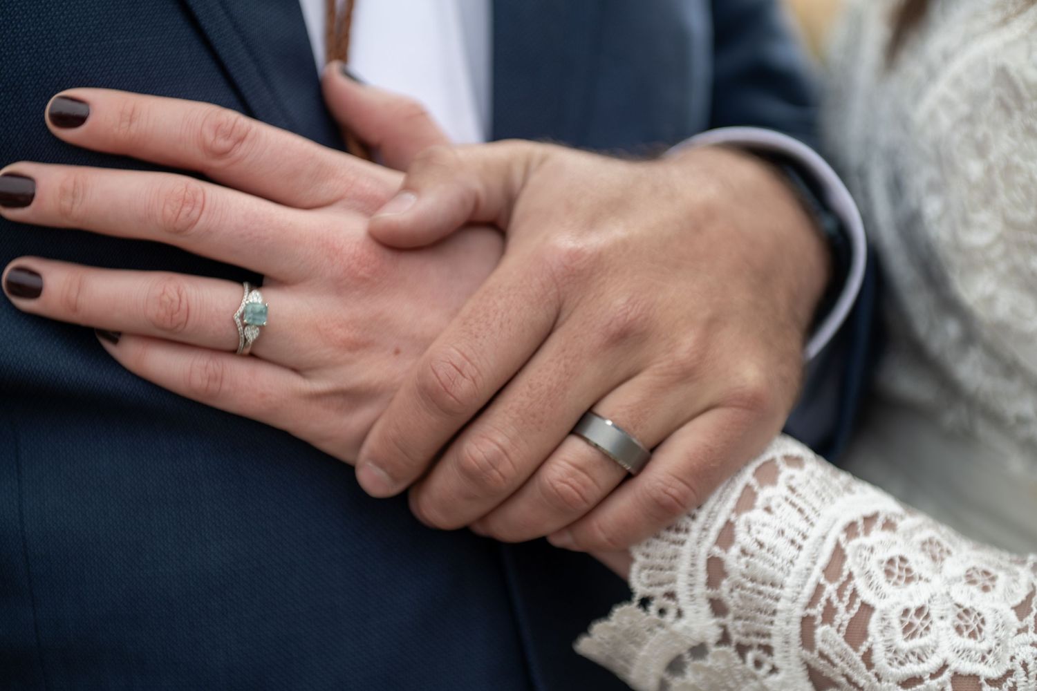 Close-up of newlywed couple's hands showing wedding rings, bride wearing lace dress, groom in blue suit.