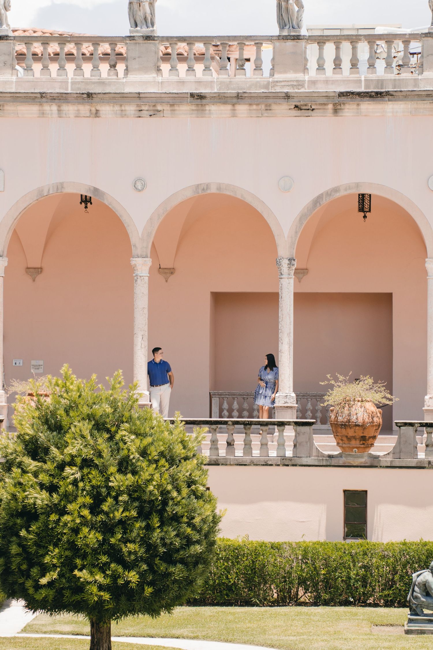 Couple standing under pink arched loggia of elegant Italian-style villa with manicured gardens.