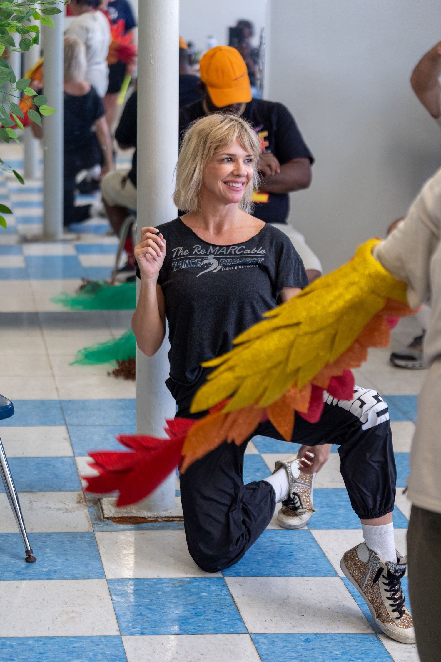 Woman kneeling on checkered floor smiling, holding colorful feathered prop with crowd in background.