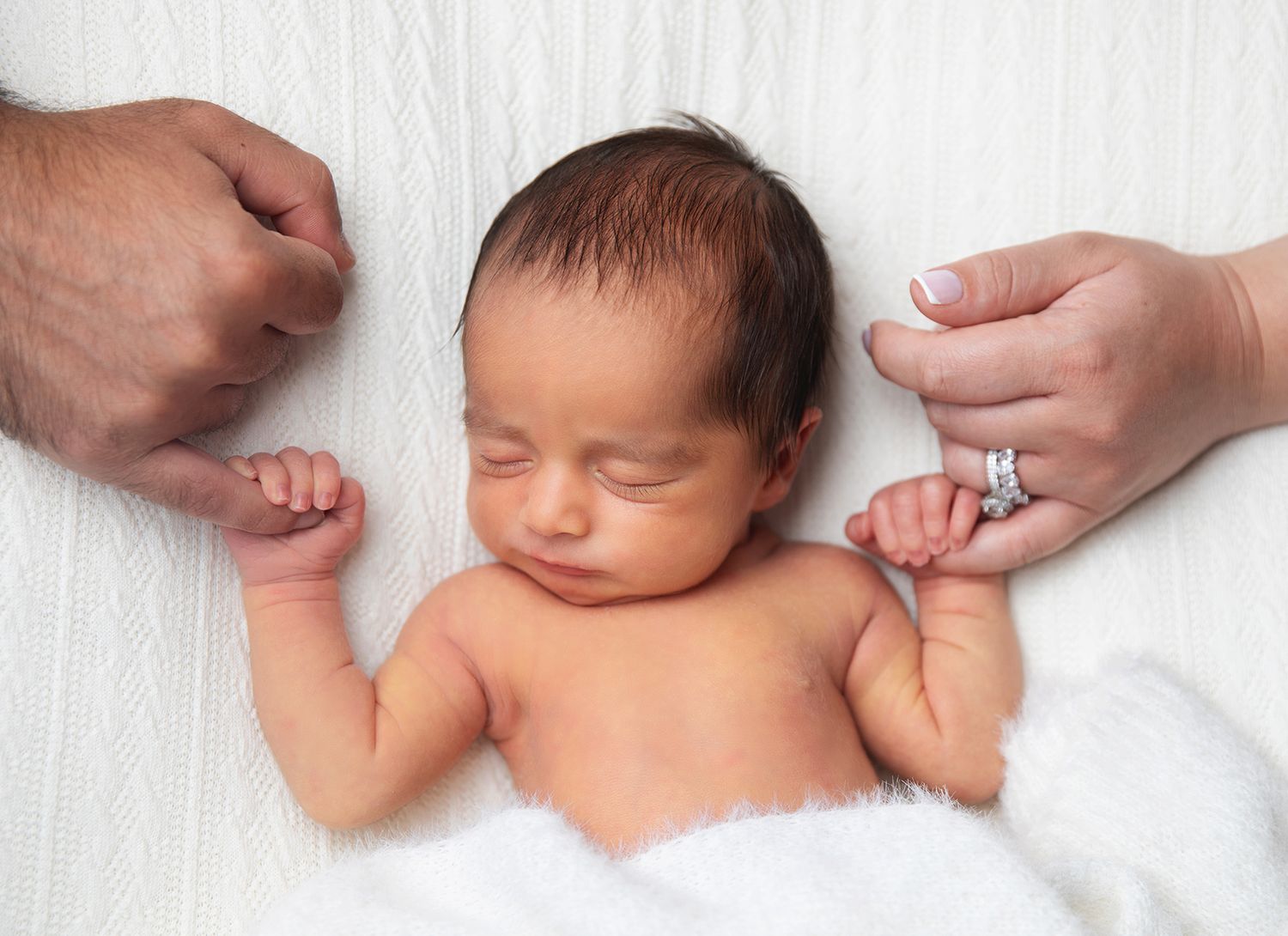 Newborn baby sleeping peacefully on white blanket while parents hold tiny hands, wearing wedding ring. During a newborn session in bexley