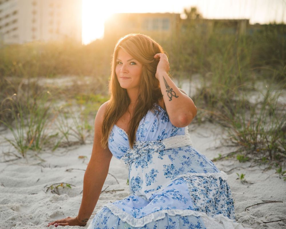 Woman in blue floral sundress sits on beach sand in warm golden sunset light.