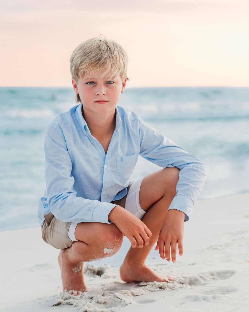 Blonde boy in light blue shirt crouches on sandy beach at dusk.