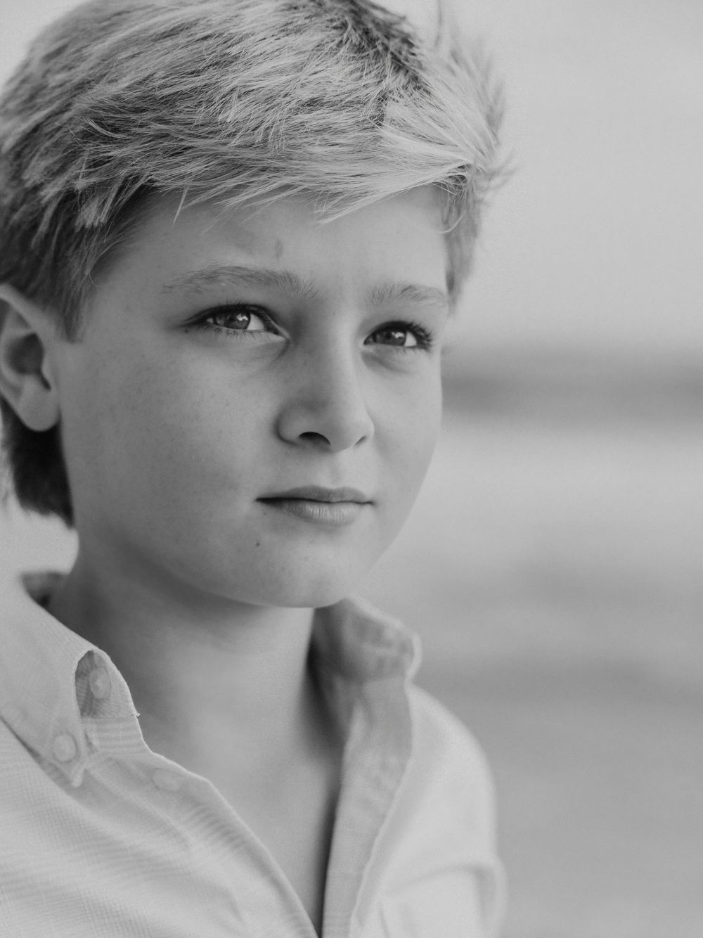 Black and white portrait of young blonde boy in collared shirt on beach.