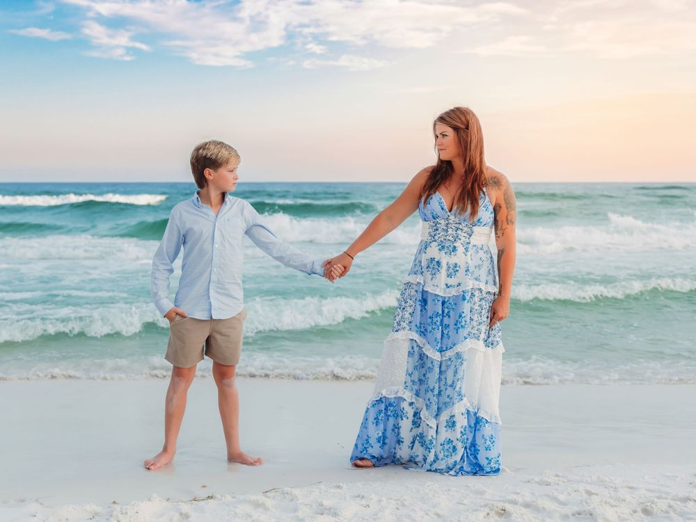 Mother and son hold hands on white sand beach at colorful sunset.