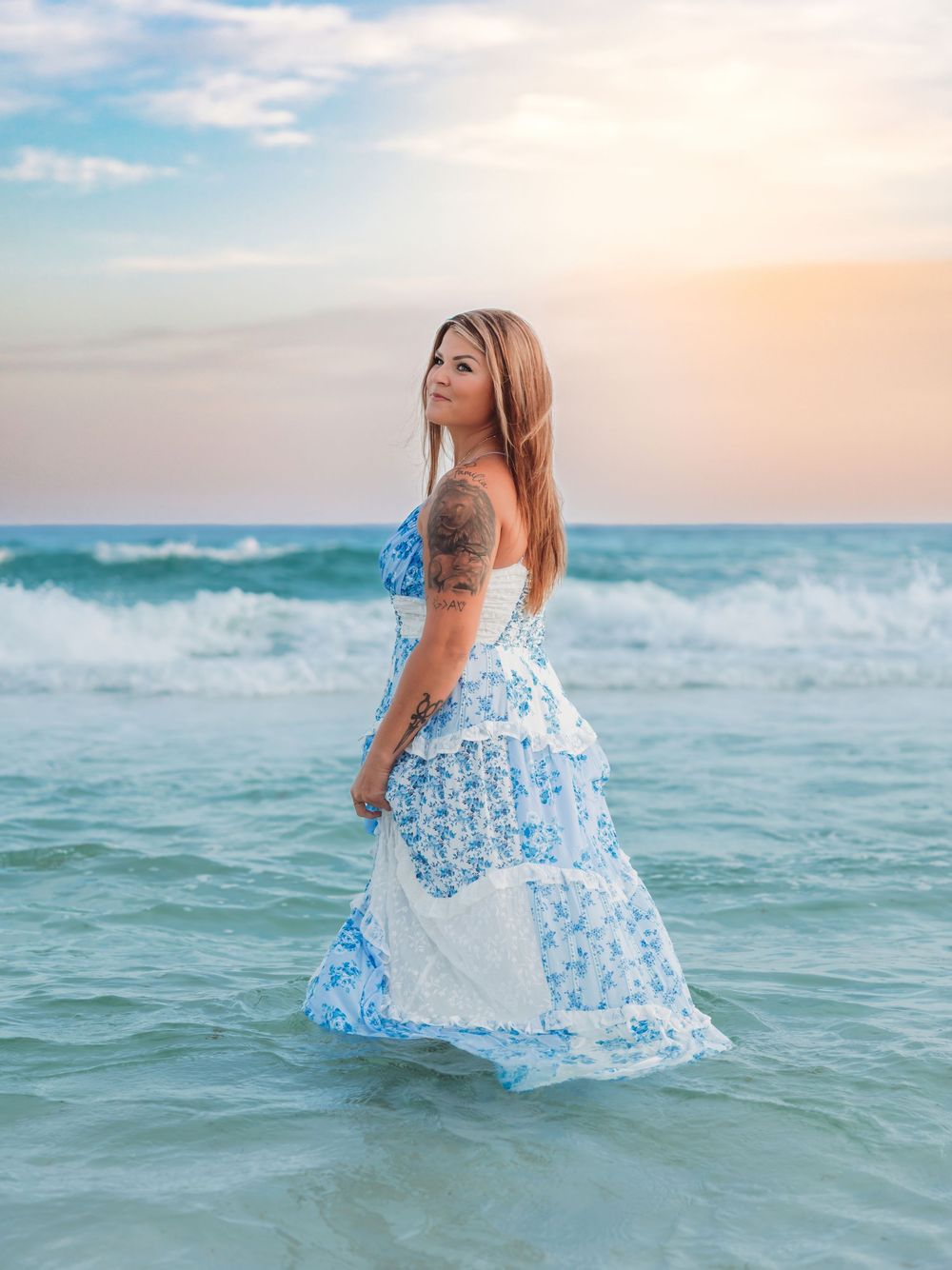 Woman in blue and white floral maxi dress stands in ocean waves at sunset.