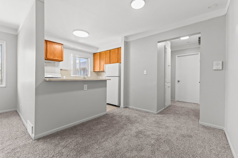 Open living area with gray carpet flows into a kitchen featuring wood cabinets and white refrigerator.