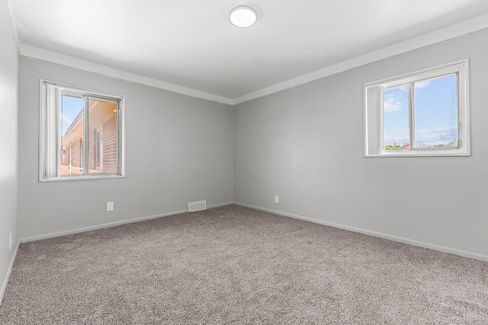 Spacious empty bedroom with gray carpet, crown molding, and two bright windows with natural light.