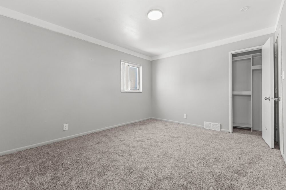 Spacious bedroom with gray carpet, crown molding, a walk-in closet, and a small window in a bright, neutral-toned room.