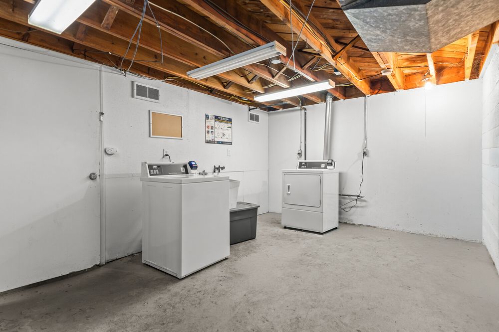 Unfinished basement laundry room with washer and dryer units, exposed wooden ceiling joists, and white painted concrete walls.