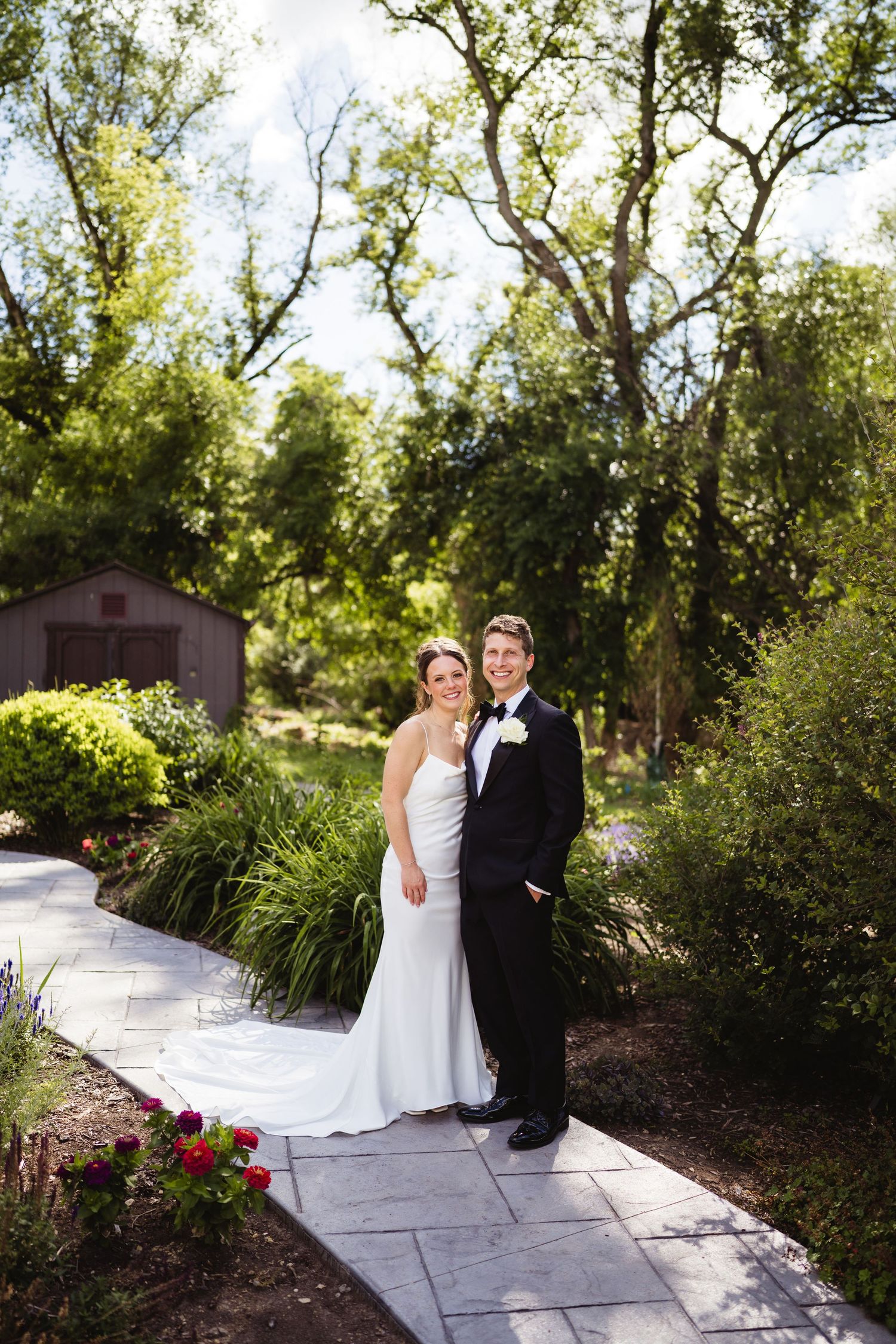 Bride and groom posing on garden path in wedding attire, surrounded by lush greenery and colorful flowers.