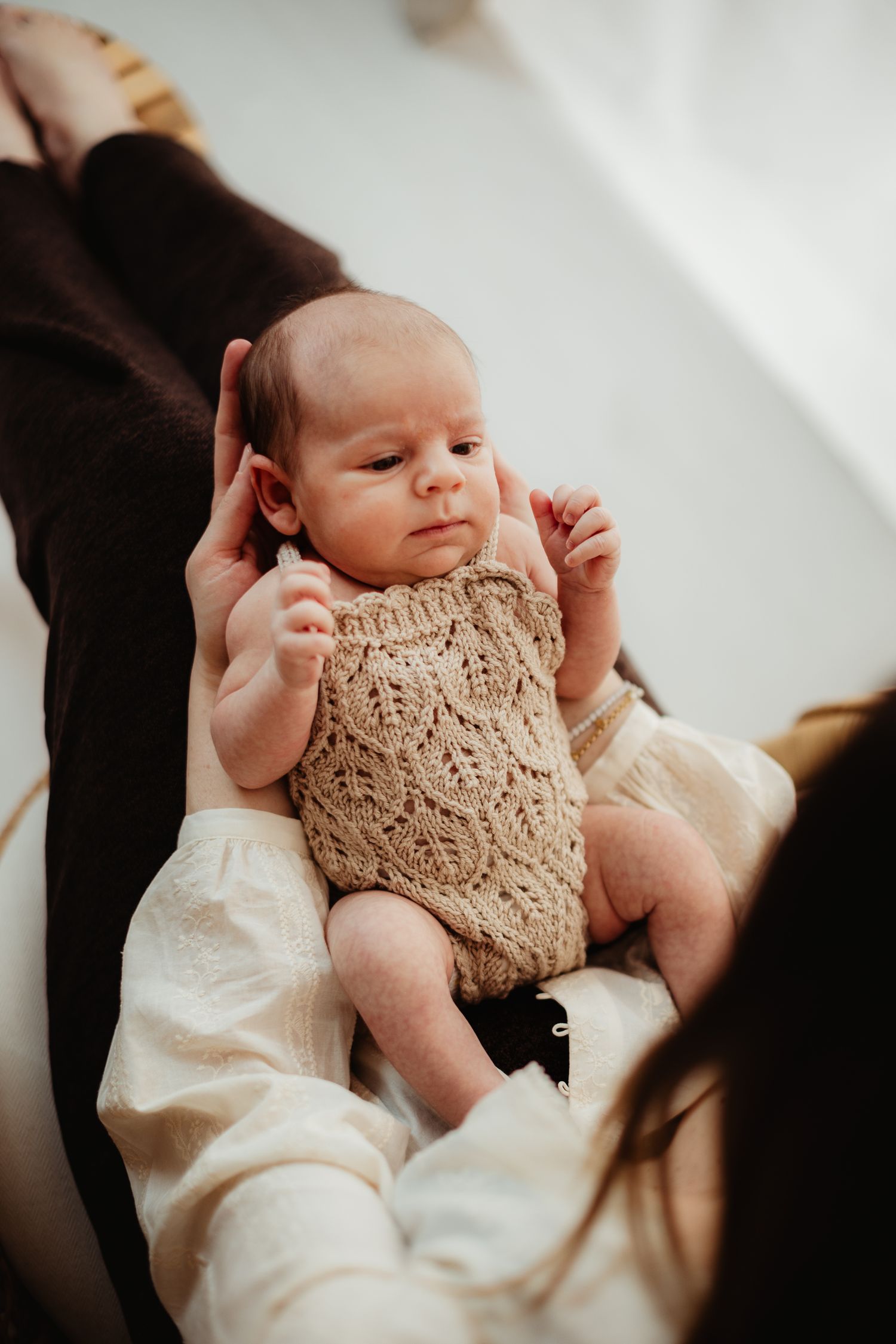 Newborn baby in knitted beige romper cradled gently in mother's arms during a soft natural light photoshoot.