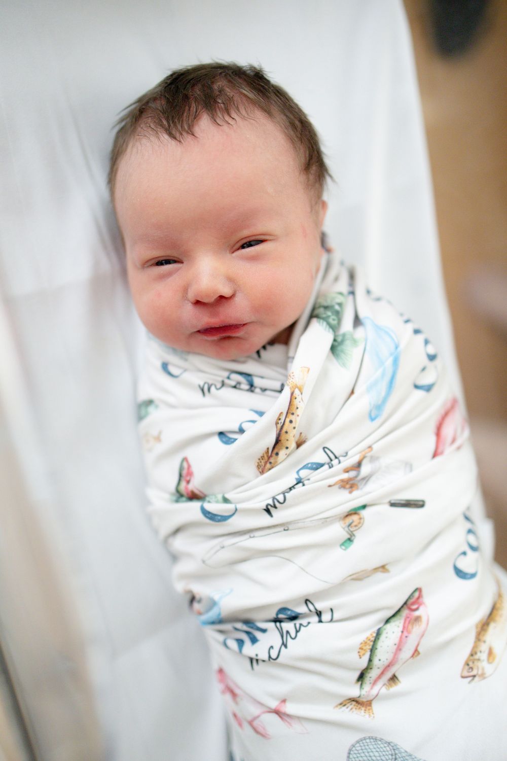 Newborn baby swaddled in a white fishing-themed blanket, lying peacefully in a hospital bassinet.