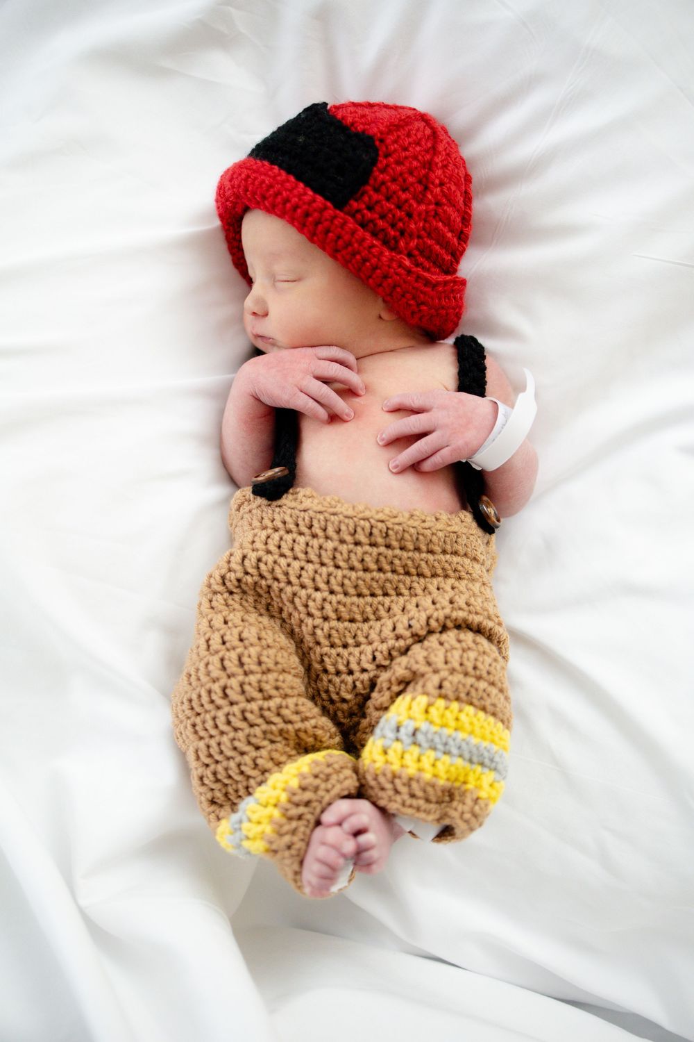 Sleeping newborn baby wearing a crocheted firefighter helmet and tan overalls costume on white bedding.