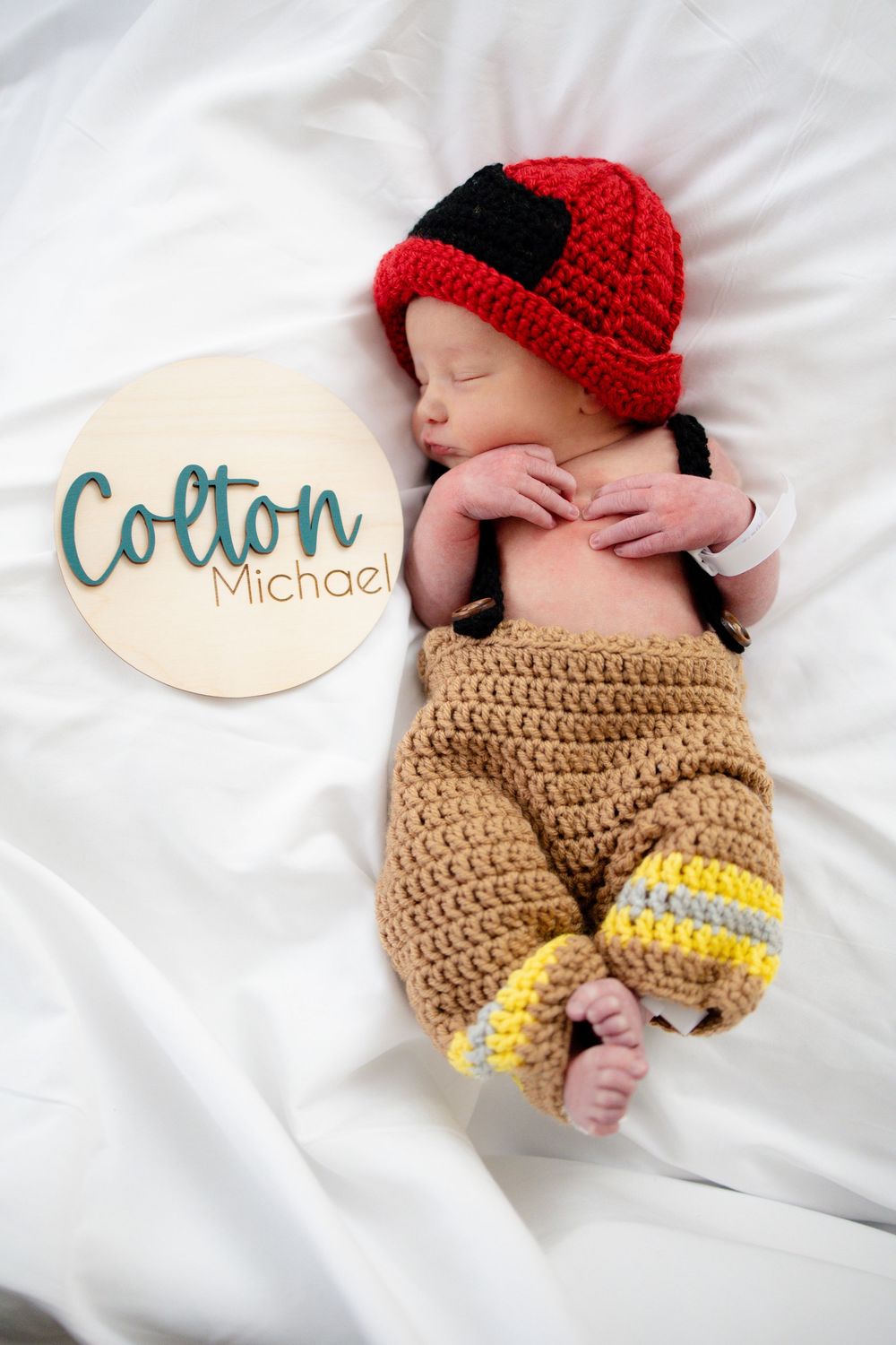 Newborn baby Colton Michael sleeping in crocheted firefighter outfit with red helmet and tan pants with yellow stripes.