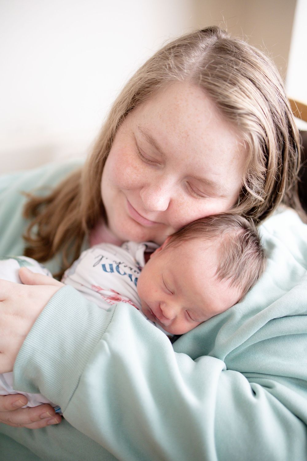 A mother with closed eyes smiles while cradling her sleeping newborn baby against her chest.