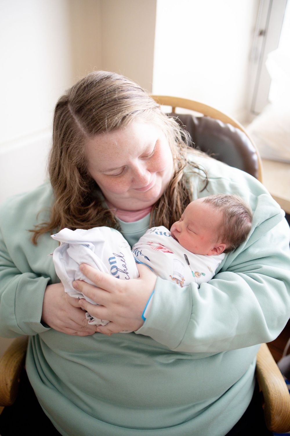Mother in mint sweatshirt tenderly holds sleeping newborn baby swaddled in white blanket while seated in chair.
