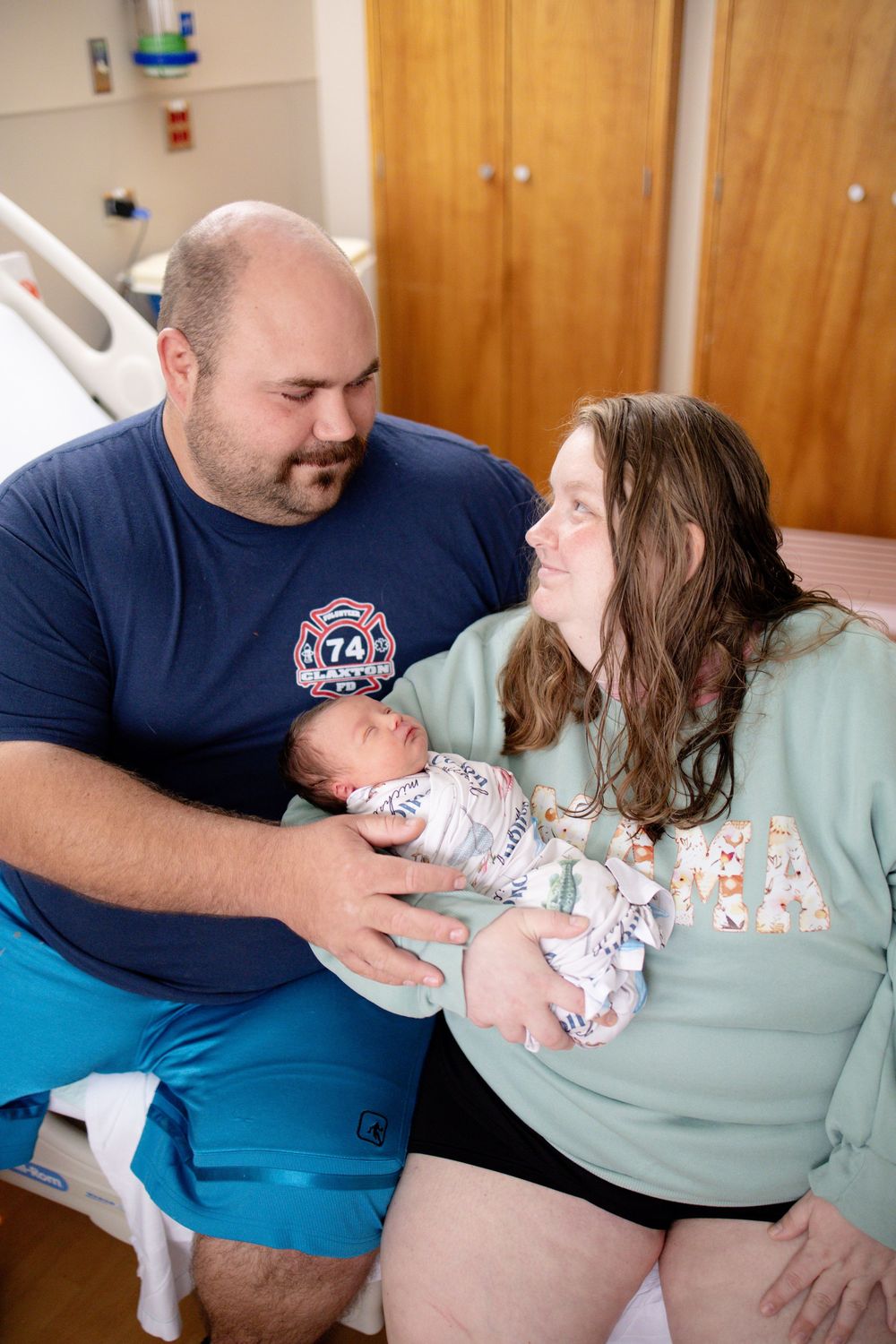 New parents in hospital room share a loving glance while holding their swaddled newborn baby together.