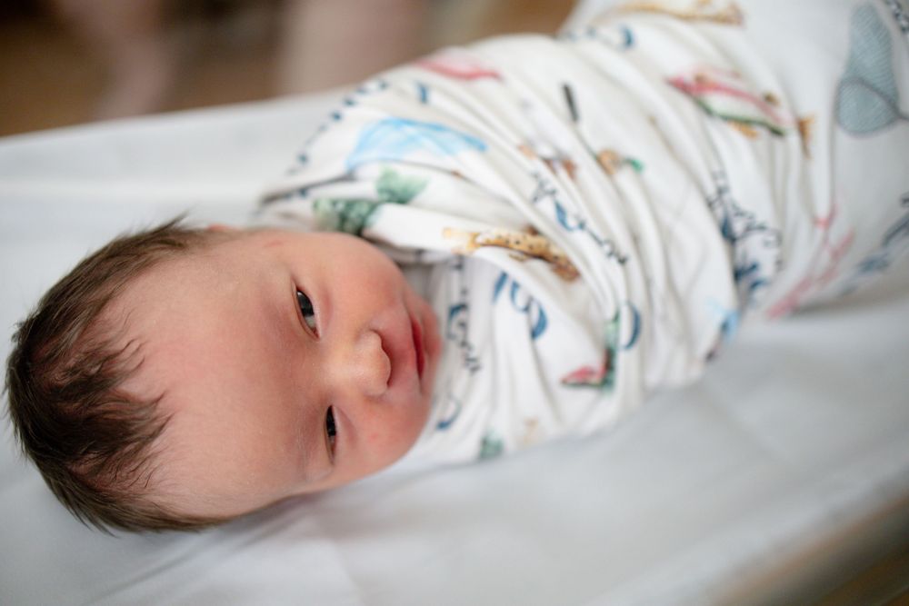 Newborn baby swaddled in a colorful printed blanket lying on a white surface in a hospital nursery.