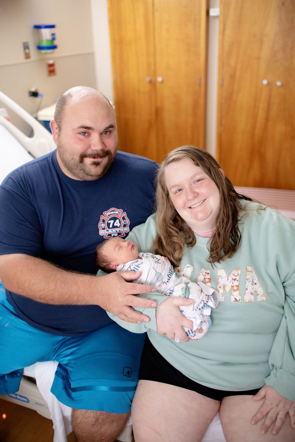 Smiling new parents pose together in hospital room holding their swaddled newborn baby after delivery.