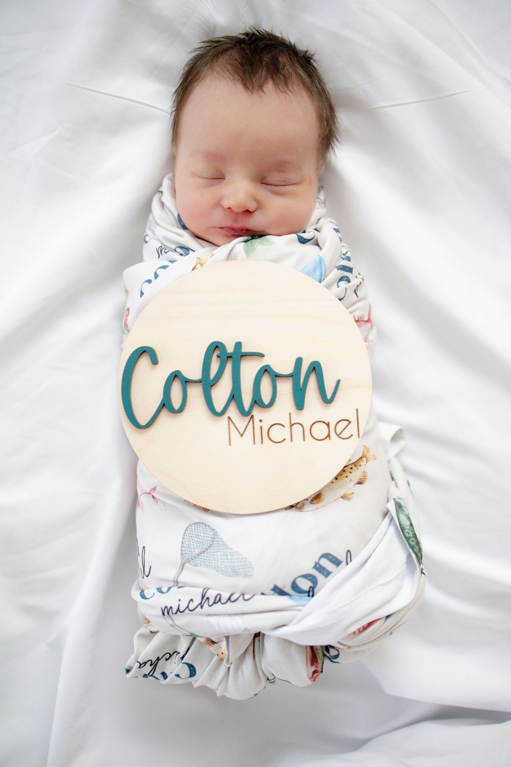 Newborn baby Colton Michael swaddled in a printed blanket with a wooden name sign on white bedding.