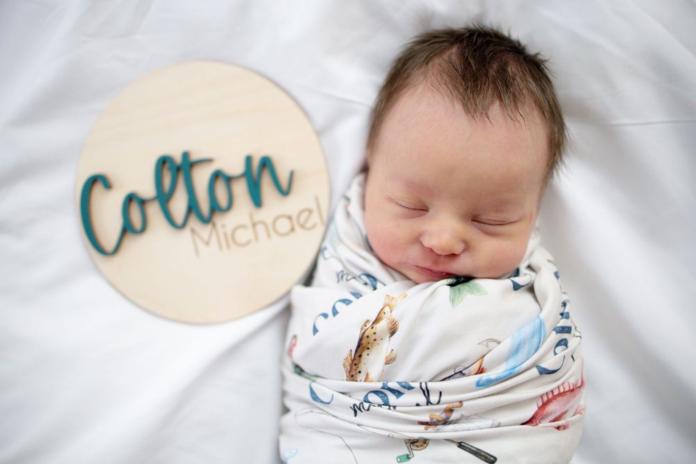 Newborn baby Colton Michael swaddled in a patterned blanket, sleeping peacefully next to a wooden name sign.