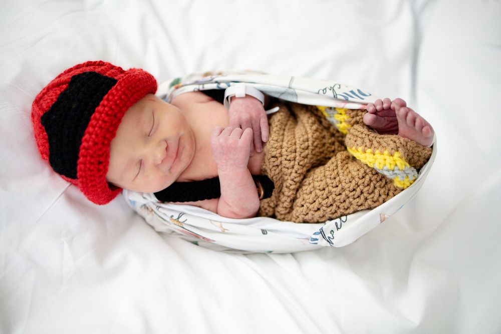 Sleeping newborn baby in a basket wearing a red and black crocheted hat and brown outfit.