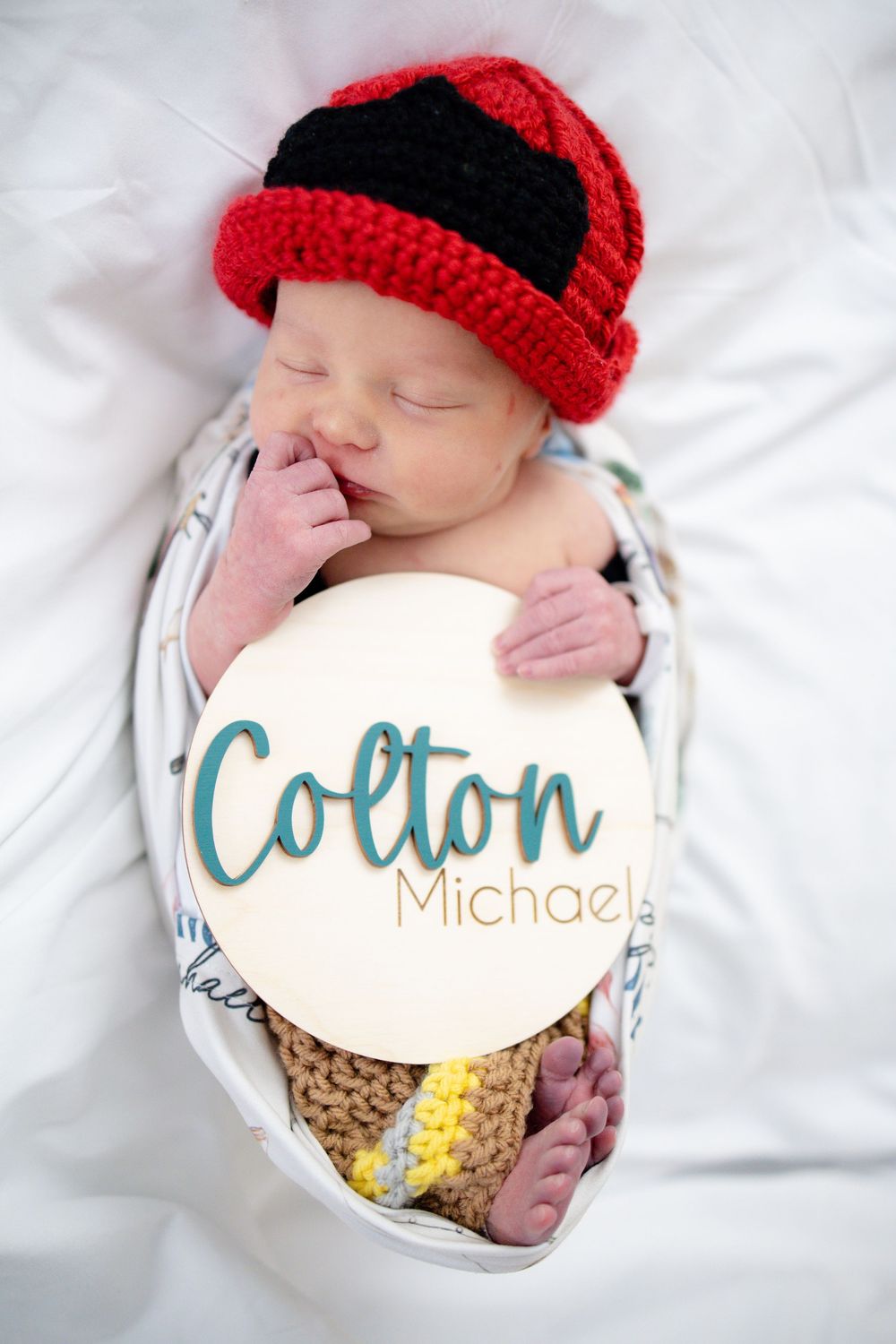 Newborn baby named Colton Michael sleeping in white swaddle wearing red and black crochet firefighter hat.